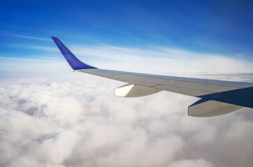Obraz premium A view from an airplane window shows a silver wing with a blue winglet flying high above a thick layer of white clouds under a clear blue sky.