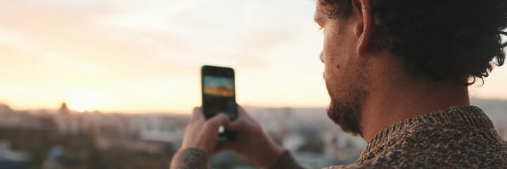 Happy guy stands on balcony at dawn, takes photo, video of beautiful views on smartphone. Panoramic. Panoramic © Andrii Nekrasov
