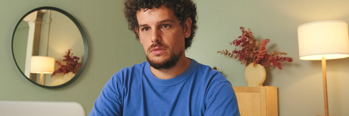 Fototapeta premium Young man with curly hair and beard using laptop in living room at table. Panoramic