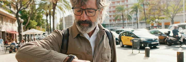 Middle-aged man stands down the street with a backpack and looks at his wristwatch. Panoramic © Andrii Nekrasov