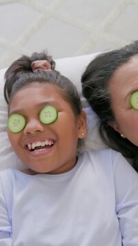 Vertical video: Laughing mom daughter on bed for fun, child adjusting cucumber, adult removing hers