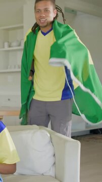 Vertical video: Entering man in yellow-blue jersey draping green flag as cape at home, cheering