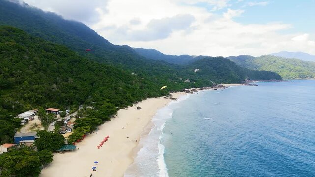Aerial view of Trindade Beach with turquoise ocean, forested mountains, paragliders and coastal village in Paraty, Brazil. Pull back and pan camera movement