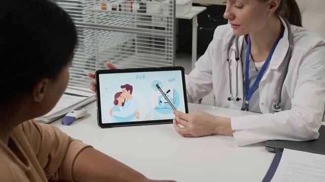 Black woman listening to positive Caucasian fertility doctor telling her about IVF cycle pointing at screen of tablet with pencil during consultation