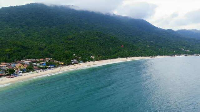Aerial view of Trindade Beach with turquoise ocean, cloud-covered mountains, paragliders and coastal village in Paraty, Brazil. Pull back and pan camera movement