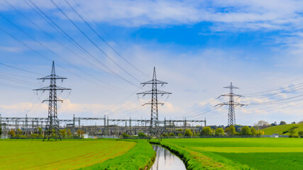 Electric towers stand near a green field and a winding stream under a blue sky with clouds in a rural setting © Fleuronica