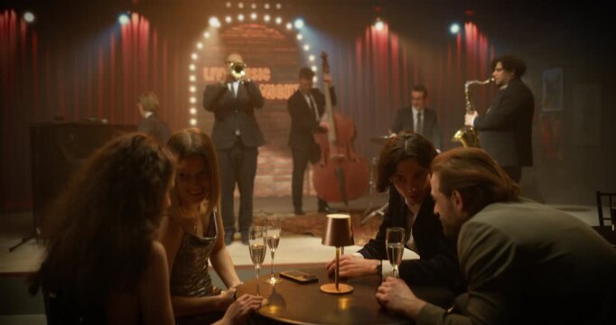 Four Adult Friends Enjoy a Fun Night at a Live Jazz Concert, Talking at Their Table, Smiling, Drinking Champagne, and Socializing While the Band Plays in a Cozy Club Setting.