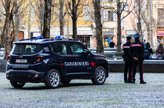 Italian military police vehicle with Carabinieri markings, blue emergency lights on roof, law enforcement officers on duty in a public square, city life background