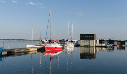  Czerwony jacht żaglowy w nowoczesnej marinie na jeziorze, spokojna woda o poranku. © Adam