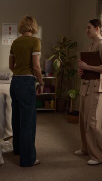 Vertical full body shot of Caucasian blonde woman and professional female massage therapist greeting each other in cozy treatment room, having little examination before therapy session