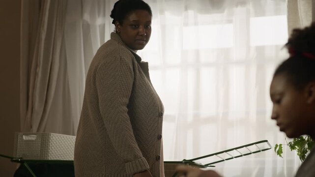Tilt down shot of African American mother finishing doing laundry and confronting teenage daughter taking away her smartphone, worried about her phone addiction