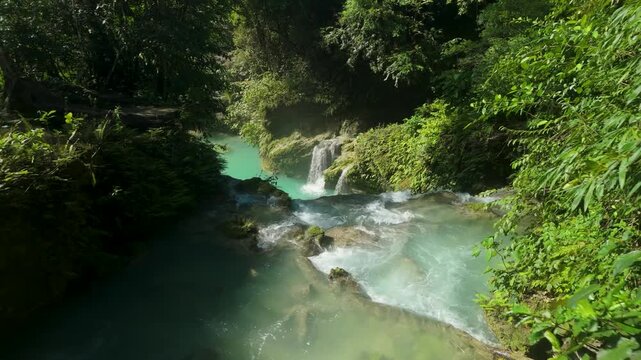 Slow aerial pan descending following Kawasan Falls river flow in Cebu Philippines revealing emerald turquoise water rushing over mossy limestone rocks small cascades lush ferns tropical canopy and dap