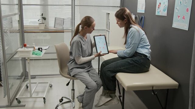 Long-haired girl pointing at digital tablet and discussing IVF steps with female fertility expert during preconception appointment in clinic