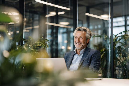 Smiling mature businessman in navy dotted blazer and open collar striped shirt, seated at laptop amid lush office plants and glass partitions