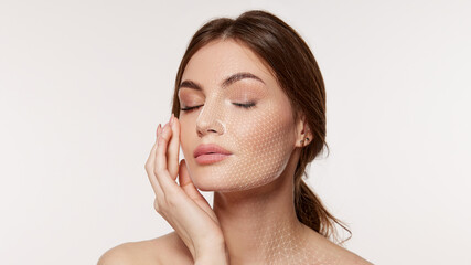 Woman with closed eyes touches cheek as fine white facial mesh covers face and neck on clean background. Ideal for skincare analysis, cosmetic treatments and beauty tech ads. © master1305