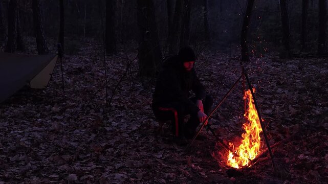 Man sitting by a campfire in a dark forest at dusk, bushcraft camping