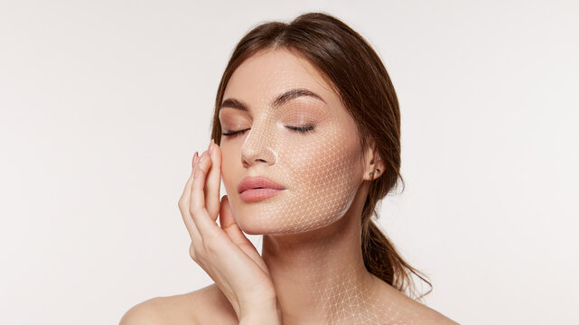 Woman with closed eyes touches cheek as fine white facial mesh covers face and neck on clean background. Ideal for skincare analysis, cosmetic treatments and beauty tech ads.