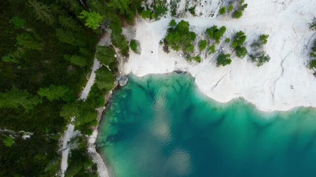 Aerial view of Lake Braies in the Dolomites, Italy, showcasing its turquoise waters adjacent to a light-colored, rocky shore. The surrounding dense forest, primarily composed of conifers like pines 