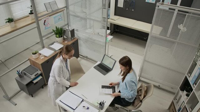 High angle view of female gynecologist entering her office, sitting down in front of young patient, having quick look at ultrasound image and inviting her to move to examination room
