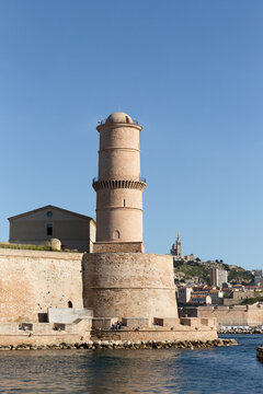 Vue sur la tour du Fanal et des remparts du fort Saint-Jean de Marseille, la mer au pied de la tour, la ville et la basilique Notre-Dame de la Garde sur la colline en arri&egrave;re-plan, grand ciel bleu