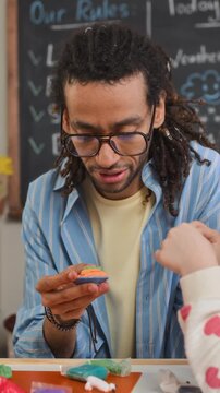 Vertical medium slowmo shot of young biracial male teacher holding handmade clay figurine sitting at table in kindergarten classroom and talking to unrecognizable child during art activity