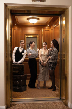 Hotel guests chat beside stacked suitcases while attendant stands near open elevator doors in warm wood interior, fitting hospitality ads, group travel and retro lifestyle promos.