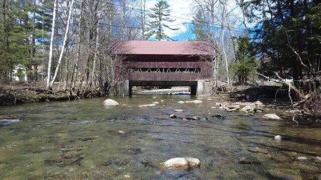 Drone pan to the left across this rocky river, focused on a historic covered bridge in Vermont