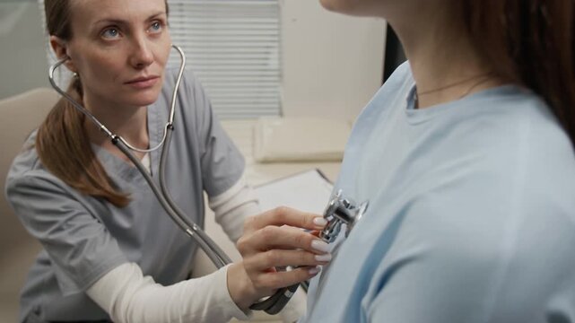 Rack focus shot of female doctor hearing lung sounds of long-haired young client using stethoscope during medical checkup