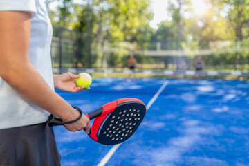 Close-up of padel racket and ball held by player before serve, with blurred doubles match in background on outdoor court, strong sports concept © Microgen
