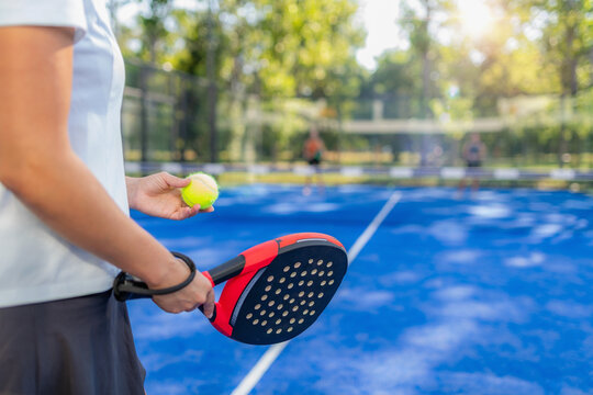 Close-up of padel racket and ball held by player before serve, with blurred doubles match in background on outdoor court, strong sports concept