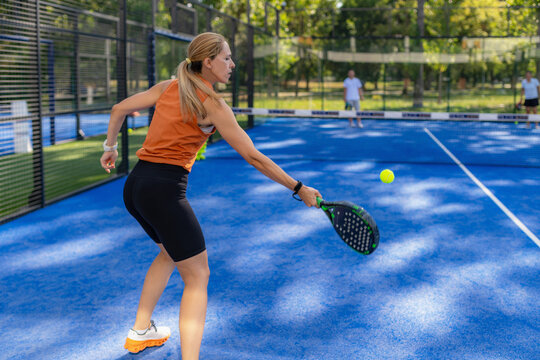 Woman hitting forehand shot during padel doubles match on outdoor court, with opponents visible across the net.