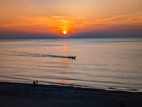 Peaceful sunset landscape with a small boat cruising on calm water and silhouettes of two people standing on the beach.