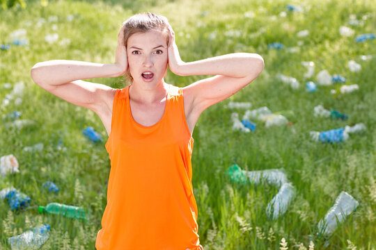 Young white woman adult holding head in shock standing in park full of plastic trash, one person reacting to pollution outdoor. Concept of environmental crisis, waste problem, nature protection