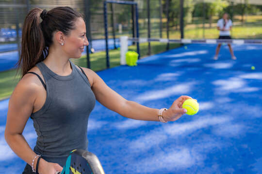 Close up of woman holding padel ball and racket before serve, with blurred opponent in background.