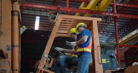 Two factory workers collaborate on a forklift, using a tablet for inventory management and strategizing logistics inside a large industrial manufacturing plant. © HarryKiiM Stock