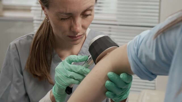 Rack focus view of young female doctor examining elbow of unrecognisable girl using dermatoscope during appointment in clinic