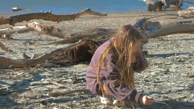 A professional static shot captures a moment of quiet observation as a young girl crouches to examine sandrocks. The textured shore of Sződliget and sun-bleached driftwood frame the scene.