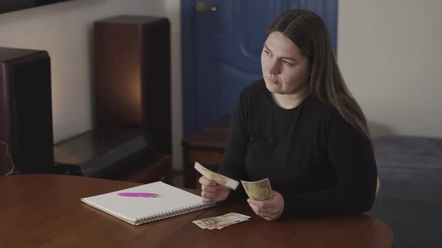Troubled woman counting cash at a table, staring blankly in financial stress, budgeting, debt concerns, or personal money problems.