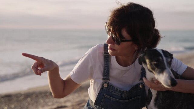 Close-up shot of a woman interacting with her dog on the beach. She gestures and talks while holding the pet, showing affection and companionship in a relaxed outdoor setting.