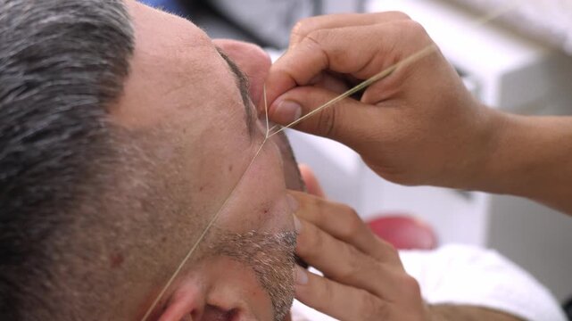 Barber shapes eyebrows using thread on a young man. The technique highlights precision, symmetry, and detailed grooming in a professional environment.