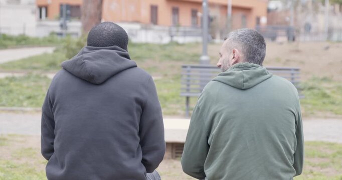 Two friends of different ethnicities talk about delicate matters in a park. From behind, one embraces the other in a supportive hug, showing care, empathy and strong friendship.