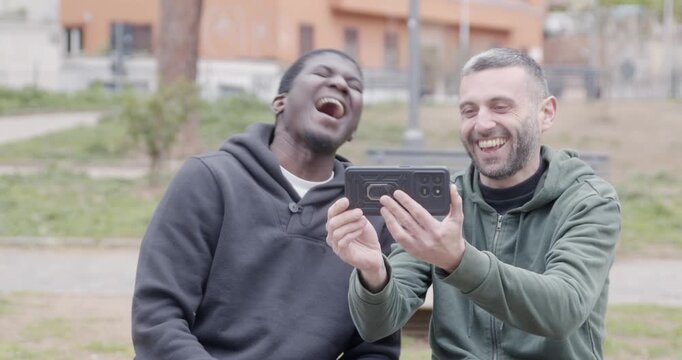 Two friends sit in a park watching a funny video on a smartphone. They burst into laughter, sharing a lighthearted moment of friendship, entertainment and digital connection.
