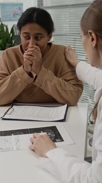 Vertical view of female gynecologist reading medical records of patient and comforting her by touching shoulder while running appointment in clinic