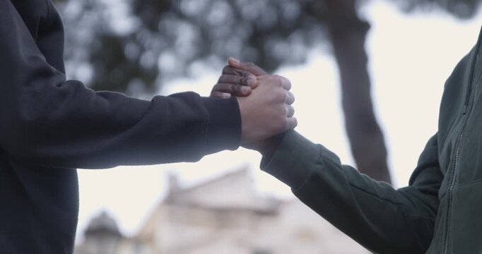 Side view close-up of two young men firmly shaking hands, one black and one white. The gesture represents unity, respect, trust and solidarity between different cultures and people.