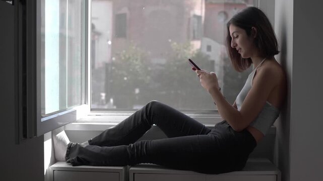 Cute dark-haired woman sits by the window smiling while texting her boyfriend. The scene conveys happiness, connection, and a light romantic moment in a cozy home setting.
