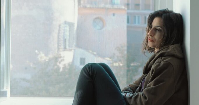 Dark-haired woman sits by the window during a cold winter day, looking sad and distant. The scene conveys loneliness, reflection, and emotional isolation