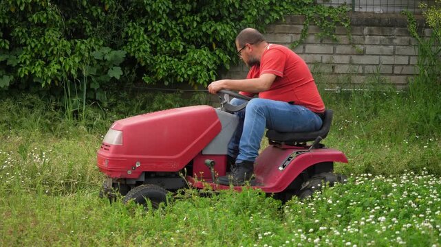 A gardener wearing a red shirt mows the lawn with a red tractor mower in a villa garden. Bright daylight enhances the scene of professional landscaping and garden care.