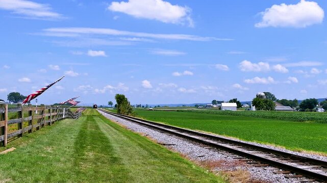 Steam Train tracks run parallel to a grassy area and fields under a blue sky. Flags wave in the breeze as the sun shines. The tracks lead toward distant buildings and trees.
