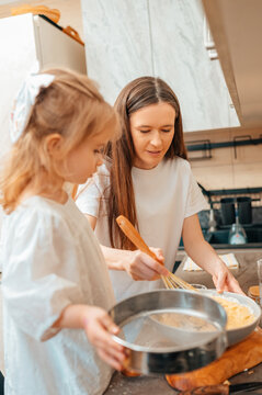 Young Caucasian Daughter Sifting Flour With Mother, Fine White Cloud Rising From Sieve, Focused Faces, Wooden