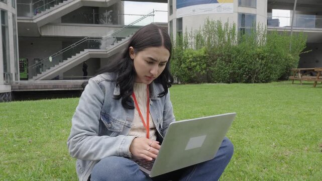 A young female student works on a laptop in a park on the university campus.